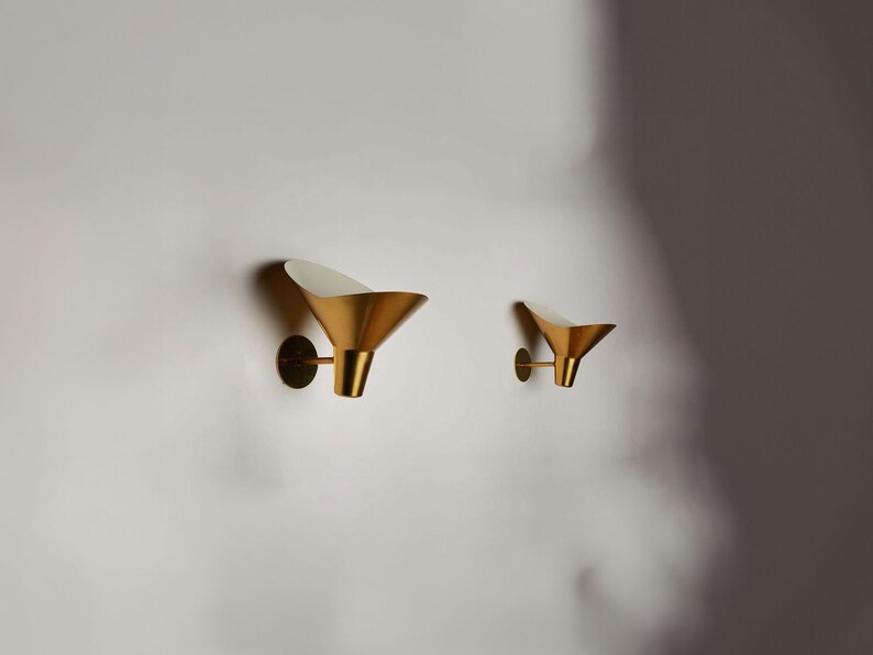 May include: Two gold wall sconces with white interior shades. The sconces are in the shape of a cone and are mounted on a white wall.