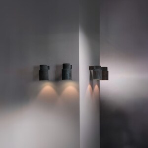 May include: Four black cylindrical wall sconces with a textured finish, mounted on a white wall. The sconces are turned on and casting a warm glow.