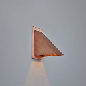 Puede incluir: Aplique de pared geométrico de color cobre. La luminaria tiene una placa trasera rectangular y una pantalla triangular que sobresale hacia afuera. La luz brilla hacia abajo, creando un brillo cálido. El aplique está montado en una pared gris claro.