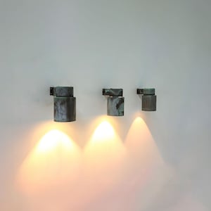 May include: Three wall-mounted, cylindrical, copper-toned light fixtures with a weathered finish. Each fixture is turned on and casting a warm, yellow light.