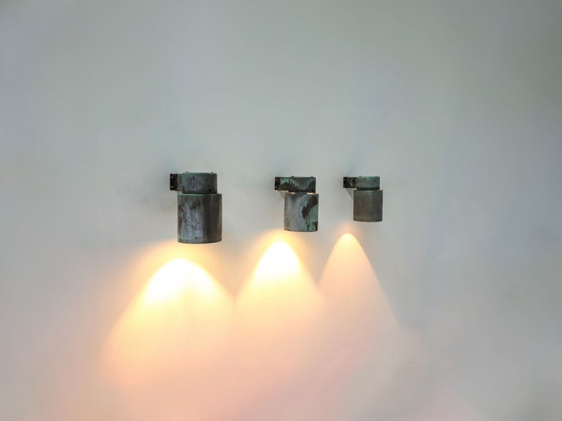May include: Three wall-mounted, cylindrical, copper-toned light fixtures with a weathered finish. Each fixture is turned on and casting a warm, yellow light.