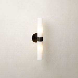 May include: A black wall sconce with two white frosted glass cylinders. The sconce has a black metal base and a black metal band around the middle of each cylinder.