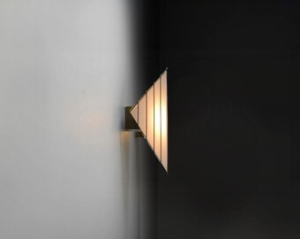 May include: A triangular wall sconce with a white frosted glass shade and a brass frame. The light is turned on and casts a warm glow.