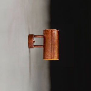 May include: A copper wall sconce with a cylindrical shade. The light is on and casting a warm glow.