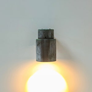 May include: A gray metal wall sconce with a round, cylindrical design. The light is on and casting a warm, yellow glow.