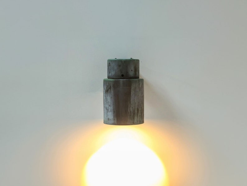 May include: A gray metal wall sconce with a round, cylindrical design. The light is on and casting a warm, yellow glow.
