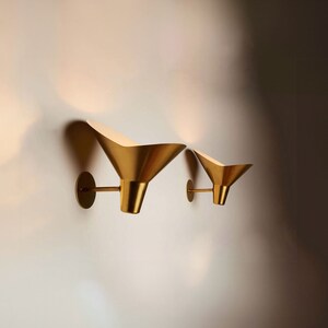 May include: Two gold-toned wall sconces with a funnel-shaped design. The sconces are mounted on a white wall.