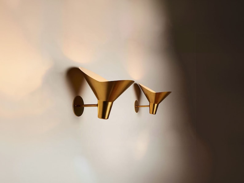 May include: Two gold-toned wall sconces with a funnel-shaped design. The sconces are mounted on a white wall.