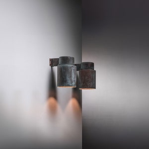 May include: Two copper wall sconces with a weathered finish, mounted on a white wall. The sconces are cylindrical in shape and have a simple design.