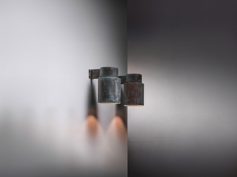 May include: Two copper wall sconces with a weathered finish, mounted on a white wall. The sconces are cylindrical in shape and have a simple design.