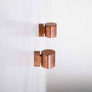 May include: Two copper wall sconces with cylindrical shades. The sconces are mounted on a white wall.