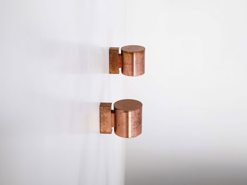 May include: Two copper wall sconces with cylindrical shades. The sconces are mounted on a white wall.