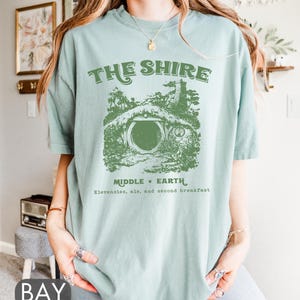 Pode incluir: Uma camiseta azul clara com um gr&aacute;fico verde de um buraco de hobbit com o texto "The Shire, Middle Earth, Elevensies, ale, and second breakfast".