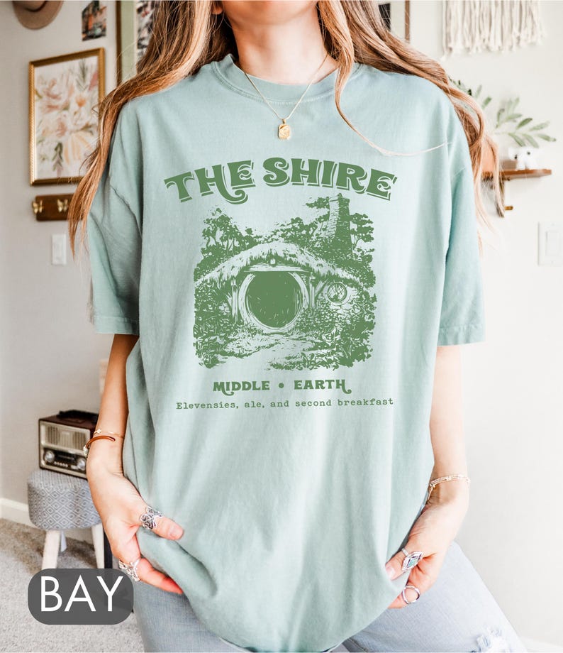 Pode incluir: Uma camiseta azul clara com um gr&aacute;fico verde de um buraco de hobbit com o texto "The Shire, Middle Earth, Elevensies, ale, and second breakfast".