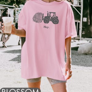 Pu&ograve; includere: Una maglietta rosa oversize con una grafica nera di un trattore che tira un fieno. Il testo "Hey." &egrave; stampato sotto la grafica.