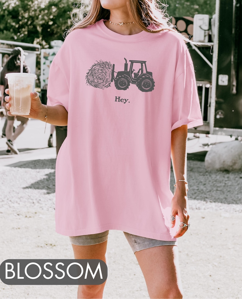 Pu&ograve; includere: Una maglietta rosa oversize con una grafica nera di un trattore che tira un fieno. Il testo "Hey." &egrave; stampato sotto la grafica.