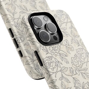 May include: Close-up of a phone case with a floral design. The case is off-white with a black outline of flowers and leaves. The camera lenses are visible, with a black border.