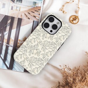 May include: A phone case with a floral design. The case is cream-colored with a pattern of black line-drawn roses and leaves. The phone case is lying on a white surface, next to a gold necklace and a magazine.