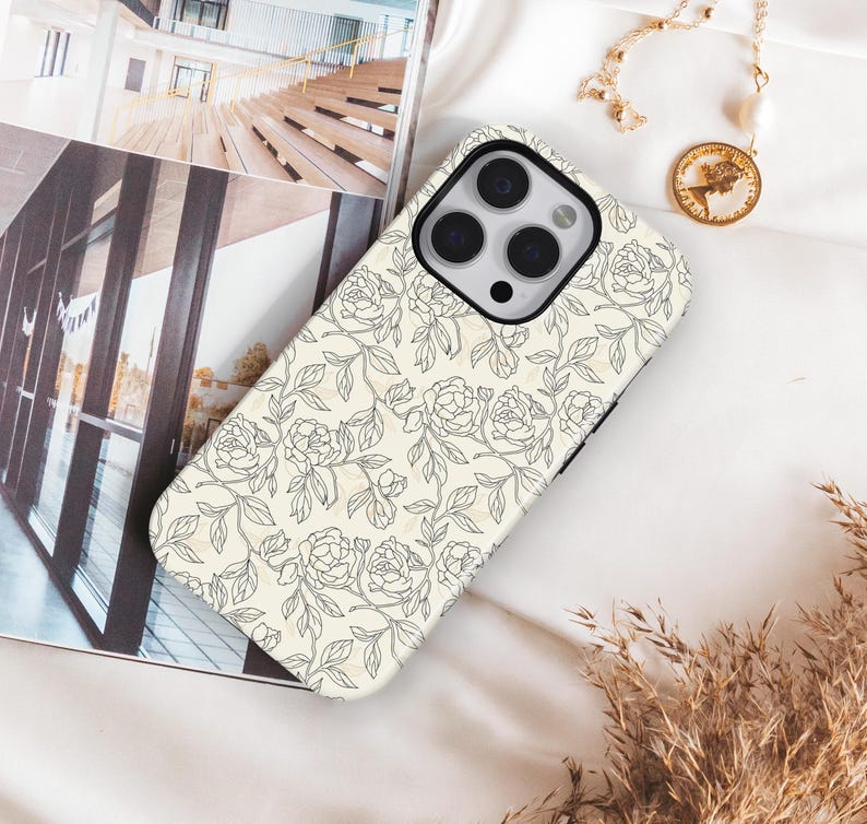 May include: A phone case with a floral design. The case is cream-colored with a pattern of black line-drawn roses and leaves. The phone case is lying on a white surface, next to a gold necklace and a magazine.