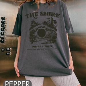 Pode incluir: Uma camiseta cinza escura com uma estampa gr&aacute;fica de uma toca de hobbit e o texto "THE SHIRE MIDDLE EARTH Elevensles, ale, and second breakfa".