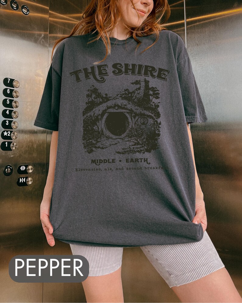 Pode incluir: Uma camiseta cinza escura com uma estampa gr&aacute;fica de uma toca de hobbit e o texto "THE SHIRE MIDDLE EARTH Elevensles, ale, and second breakfa".