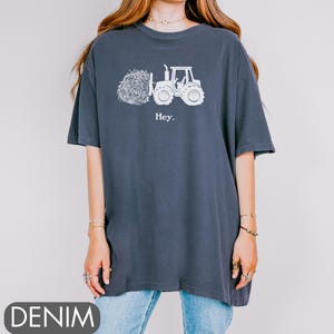 Pu&ograve; includere: Una maglietta oversize blu navy con una grafica bianca di un trattore che trasporta una balla di fieno. Il testo "Hey." &egrave; stampato sotto la grafica.