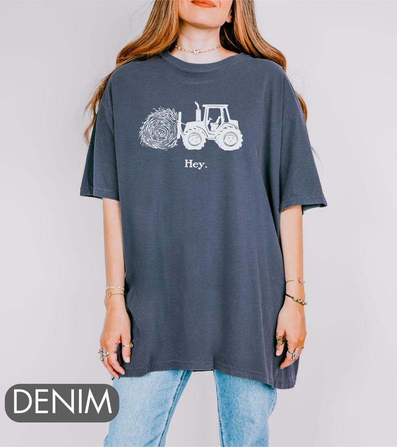 Pu&ograve; includere: Una maglietta oversize blu navy con una grafica bianca di un trattore che trasporta una balla di fieno. Il testo "Hey." &egrave; stampato sotto la grafica.