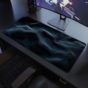 May include: A large, rectangular mouse pad with a topographic map design in shades of teal and black. The pad is placed on a dark desk, with a computer monitor and speaker visible in the background. The design features contour lines.