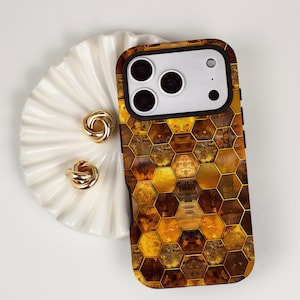 Puede incluir: Una funda de teléfono con un patrón de panal en tonos marrones y dorados. Junto a ella, un plato blanco en forma de concha con dos pendientes dorados en forma de nudo. La funda tiene un borde negro y un módulo de cámara.