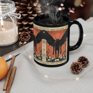 May include: Black ceramic mug with a white and orange illustration of a creature with large wings and red eyes. The creature is standing in a forest setting with a red sun in the background.
