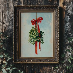 May include: A framed print featuring a sprig of mistletoe tied with a red ribbon and bow. The artwork has a light blue background and is set in an ornate, dark gold frame. The image is against a dark wood background.