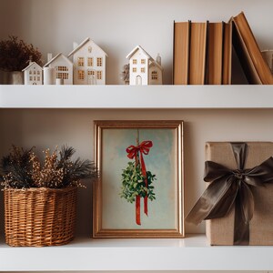 May include: A shelf displays a framed mistletoe print with a red bow, a wrapped gift with a brown satin ribbon, and a woven basket with decorative branches. Miniature white houses and stacked books complete the holiday-themed decor.