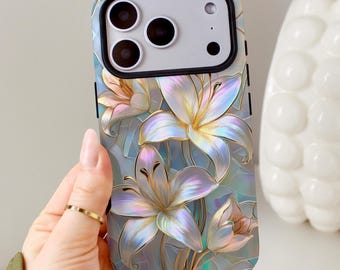 Può includere: Una custodia per smartphone con un design floreale iridescente a più strati con fiori bianchi e viola chiaro simili a gigli. I petali hanno contorni dorati e una finitura perlata su uno sfondo tenue color azzurro e grigio.