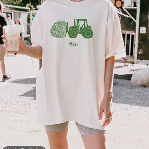 Pu&ograve; includere: Una maglietta bianca oversize con una grafica di un trattore verde e una balla di fieno. Il testo "Hey." &egrave; stampato sotto la grafica. La maglietta &egrave; indossata da una persona.