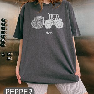Pu&ograve; includere: Una maglietta oversize di colore grigio scuro con una grafica bianca di un trattore che tira una balla di fieno. Il testo "Hey." &egrave; stampato sotto la grafica.
