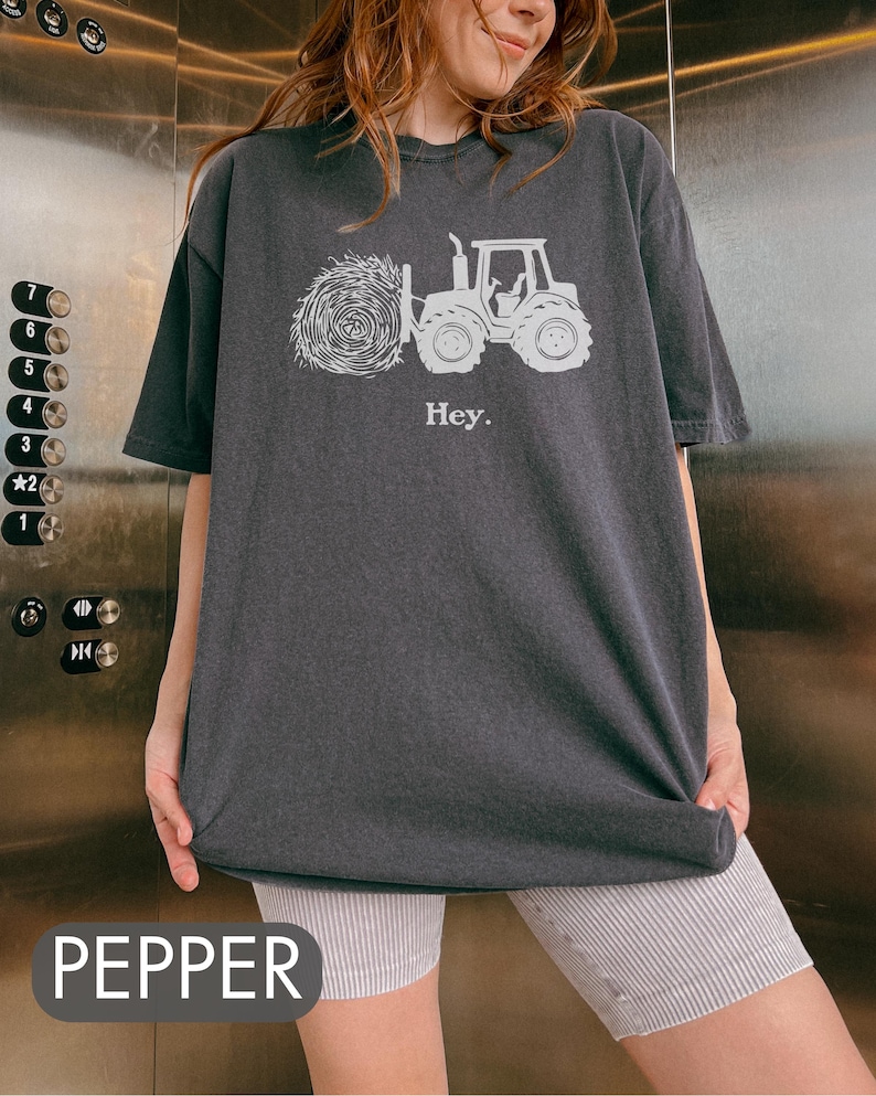 Pu&ograve; includere: Una maglietta oversize di colore grigio scuro con una grafica bianca di un trattore che tira una balla di fieno. Il testo "Hey." &egrave; stampato sotto la grafica.