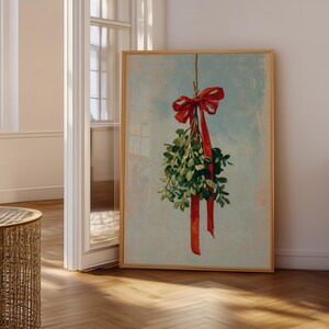 May include: A framed print featuring a sprig of mistletoe with a red bow and ribbon against a light blue background. The artwork is displayed in a room with wooden floors and a window.