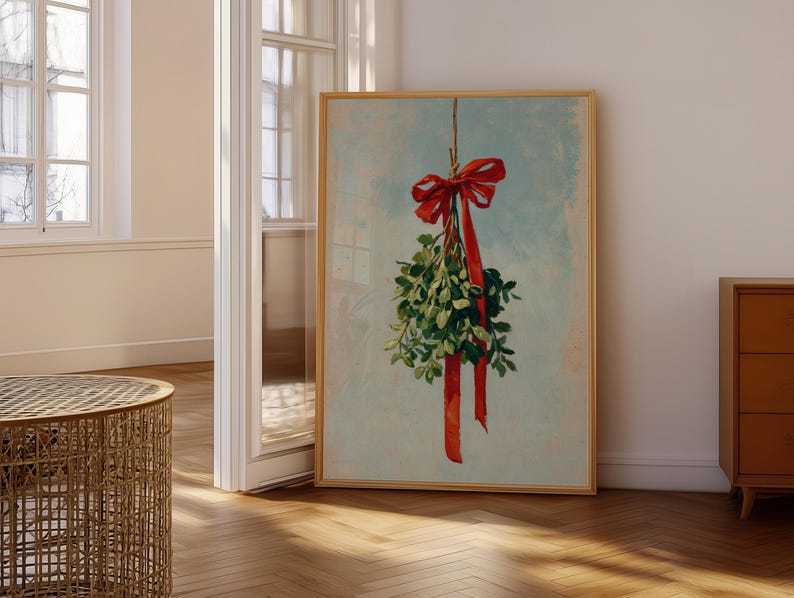 May include: A framed print featuring a sprig of mistletoe with a red bow and ribbon against a light blue background. The artwork is displayed in a room with wooden floors and a window.