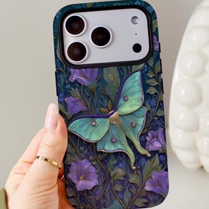 Puede incluir: Una funda rectangular para smartphone con fondo azul oscuro y un patrón de flores moradas y follaje verde. Una polilla grande de color verde claro con detalles morados y pequeñas marcas circulares está centrada en la parte posterior.