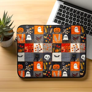 Peut inclure: Housse d'ordinateur portable noire avec un motif patchwork sur le thème d'Halloween. Le motif comprend des citrouilles oranges, des fantômes blancs, des chats noirs, des crânes et des motifs floraux. La housse est sur une surface en bois.