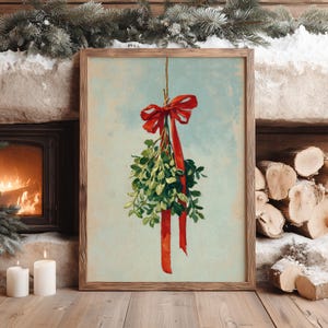 May include: A framed print of mistletoe with a red bow and ribbon. The artwork features green mistletoe leaves and a red bow against a light blue background. The frame is a light brown color. The image is set against a rustic background with a fireplace and candles.