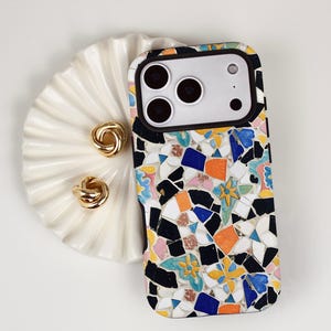 Puede incluir: Una funda de teléfono inteligente con un diseño de mosaico colorido, con fragmentos negros, blancos, azules, naranjas y amarillos. También hay dos pendientes dorados con forma de nudo y un plato blanco con forma de concha.