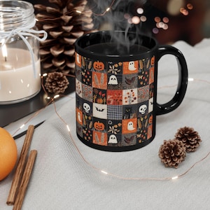 May include: A black ceramic mug featuring a repeating patchwork pattern of Halloween-themed illustrations including orange pumpkins, white ghosts, black cats, skulls, bats, and autumn foliage in shades of orange, black, gray, and white.