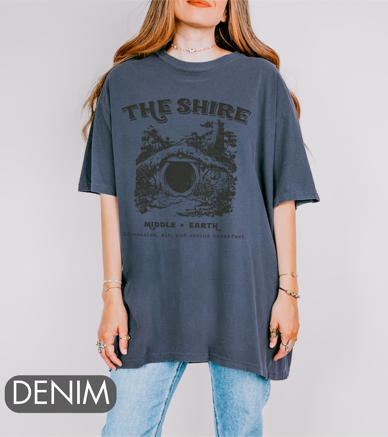 Pode incluir: Uma camiseta azul marinho com um gr&aacute;fico de uma toca de hobbit e o texto "The Shire, Middle-Earth, Second Breakfast, Ale, and Lembas".