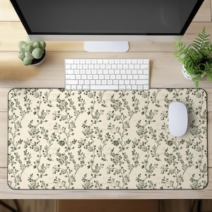 Puede incluir: Alfombrilla de ratón rectangular con fondo crema y un patrón floral repetido en tonos verdes. Un ratón y un teclado de ordenador blancos están sobre el escritorio. El escritorio es de madera clara.