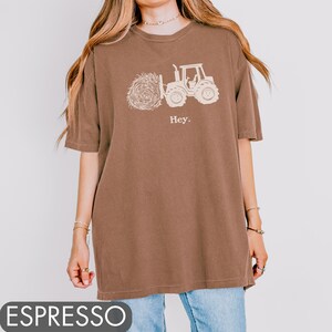 Pu&ograve; includere: Una maglietta oversize marrone con una grafica bianca di un trattore che trasporta una balla di fieno. Il testo "Hey." &egrave; stampato sotto la grafica.