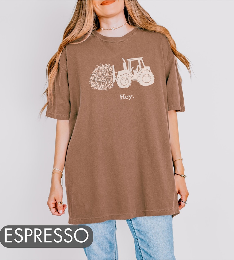 Pu&ograve; includere: Una maglietta oversize marrone con una grafica bianca di un trattore che trasporta una balla di fieno. Il testo "Hey." &egrave; stampato sotto la grafica.