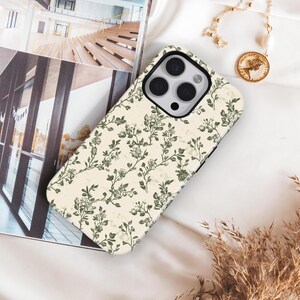 Puede incluir: Funda de tel&eacute;fono crema con un dise&ntilde;o floral verde. La funda est&aacute; colocada sobre una superficie blanca, junto a un collar dorado con un colgante de moneda y una revista.
