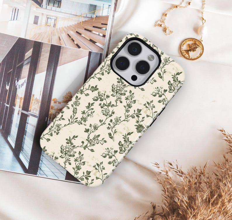Puede incluir: Funda de tel&eacute;fono crema con un dise&ntilde;o floral verde. La funda est&aacute; colocada sobre una superficie blanca, junto a un collar dorado con un colgante de moneda y una revista.