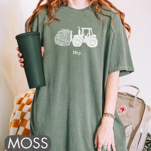 Pu&ograve; includere: Una maglietta oversize verde con una grafica bianca di un trattore che trasporta una balla di fieno. Il testo "Hey." &egrave; stampato sotto la grafica.